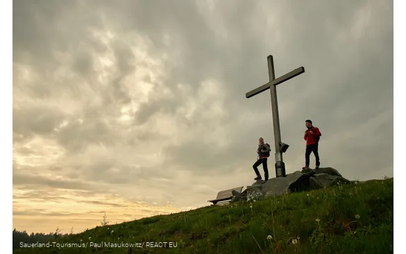 Zwei Wandernde auf einem Berggipfel neben einem Gipfelkreuz