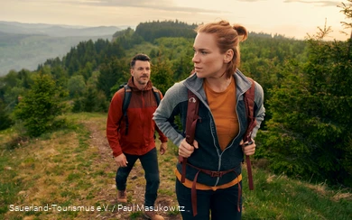 Ein junges Paar wandert einen Berg hinauf. Im Hintergrund ist die üppige, grüne Landschaft zu sehen.