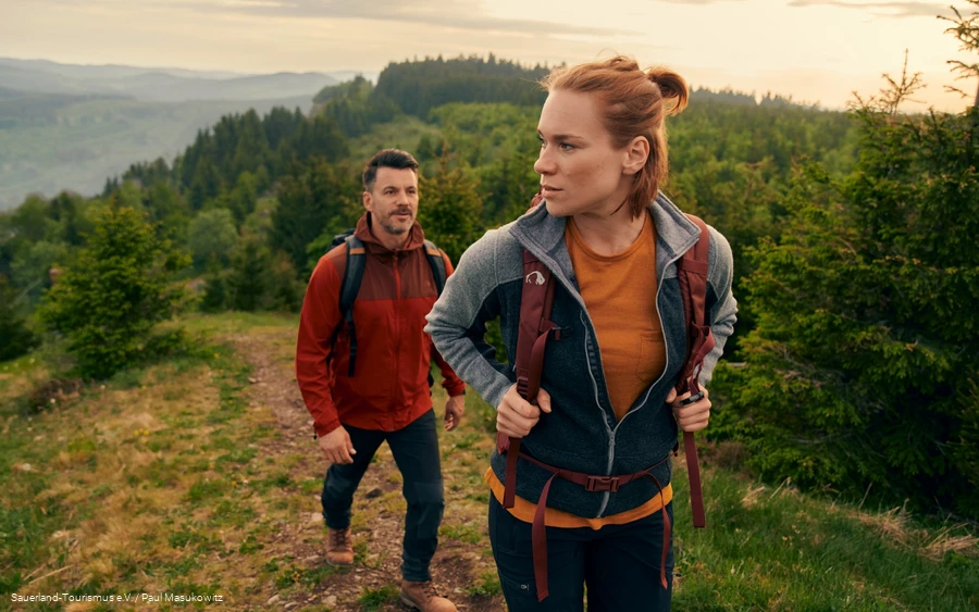 Ein junges Paar wandert einen Berg hinauf. Im Hintergrund ist die üppige, grüne Landschaft zu sehen.